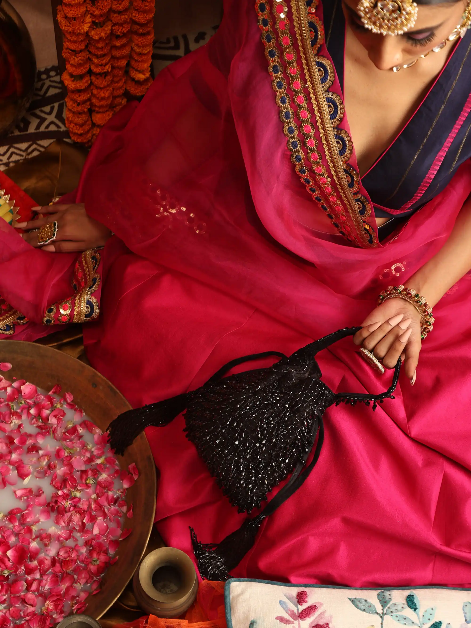Woman in a vibrant pink saree with intricate patterns, holding a black beaded bag, surrounded by decorative elements.