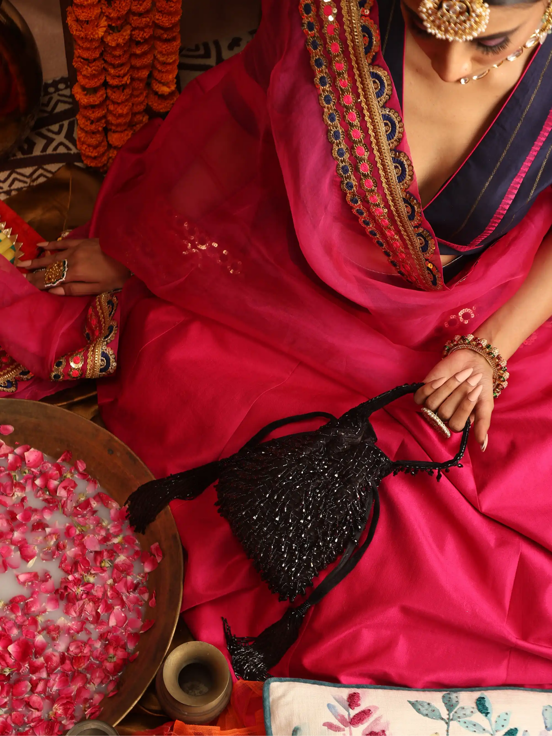 Woman in a vibrant pink saree with intricate patterns, holding a black beaded bag, surrounded by decorative elements.