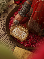 Gold clutch with intricate designs held by a hand on a decorative surface with red flowers.