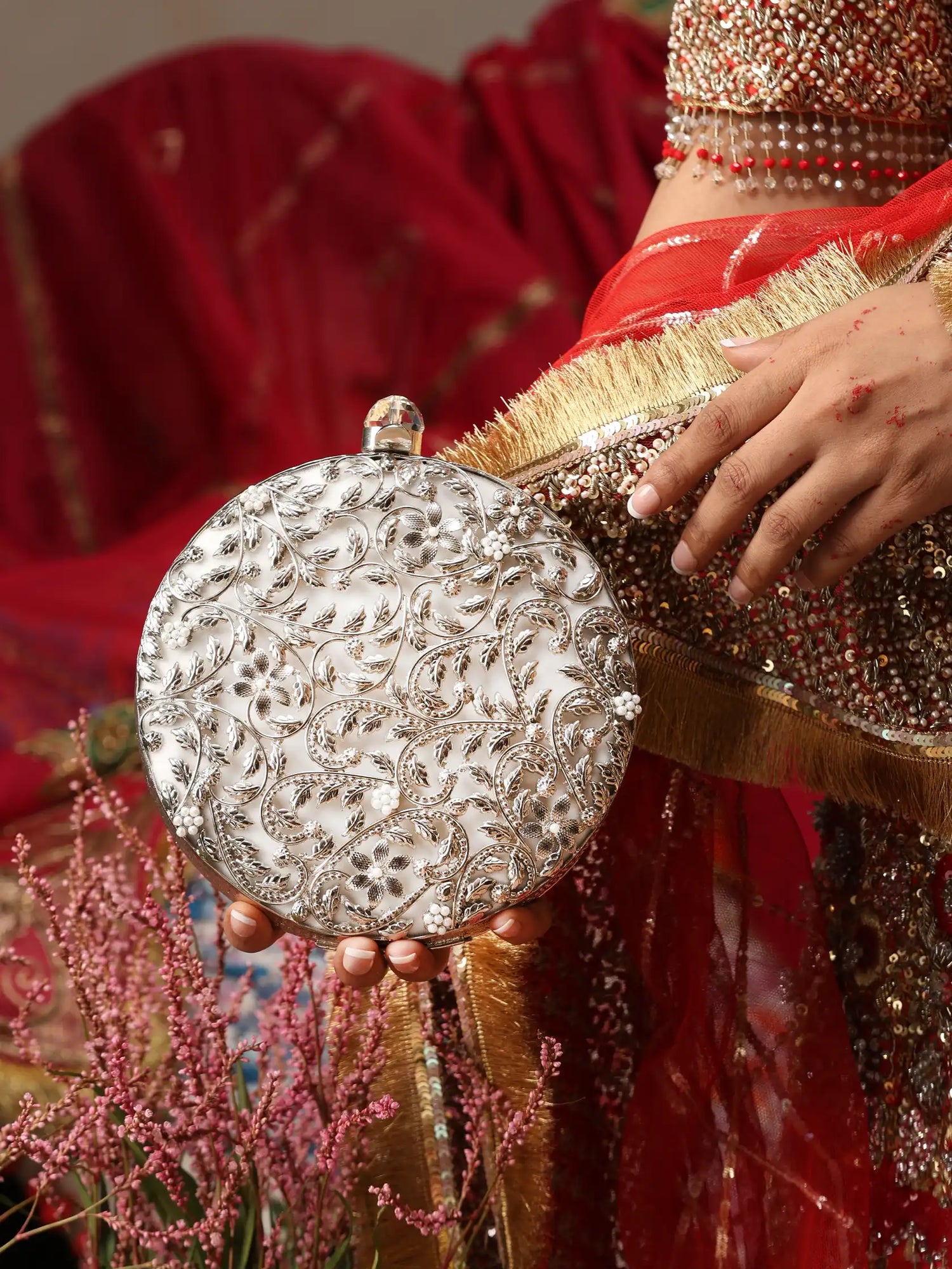 Person holding a silver clutch with intricate designs, wearing a red saree with gold border.