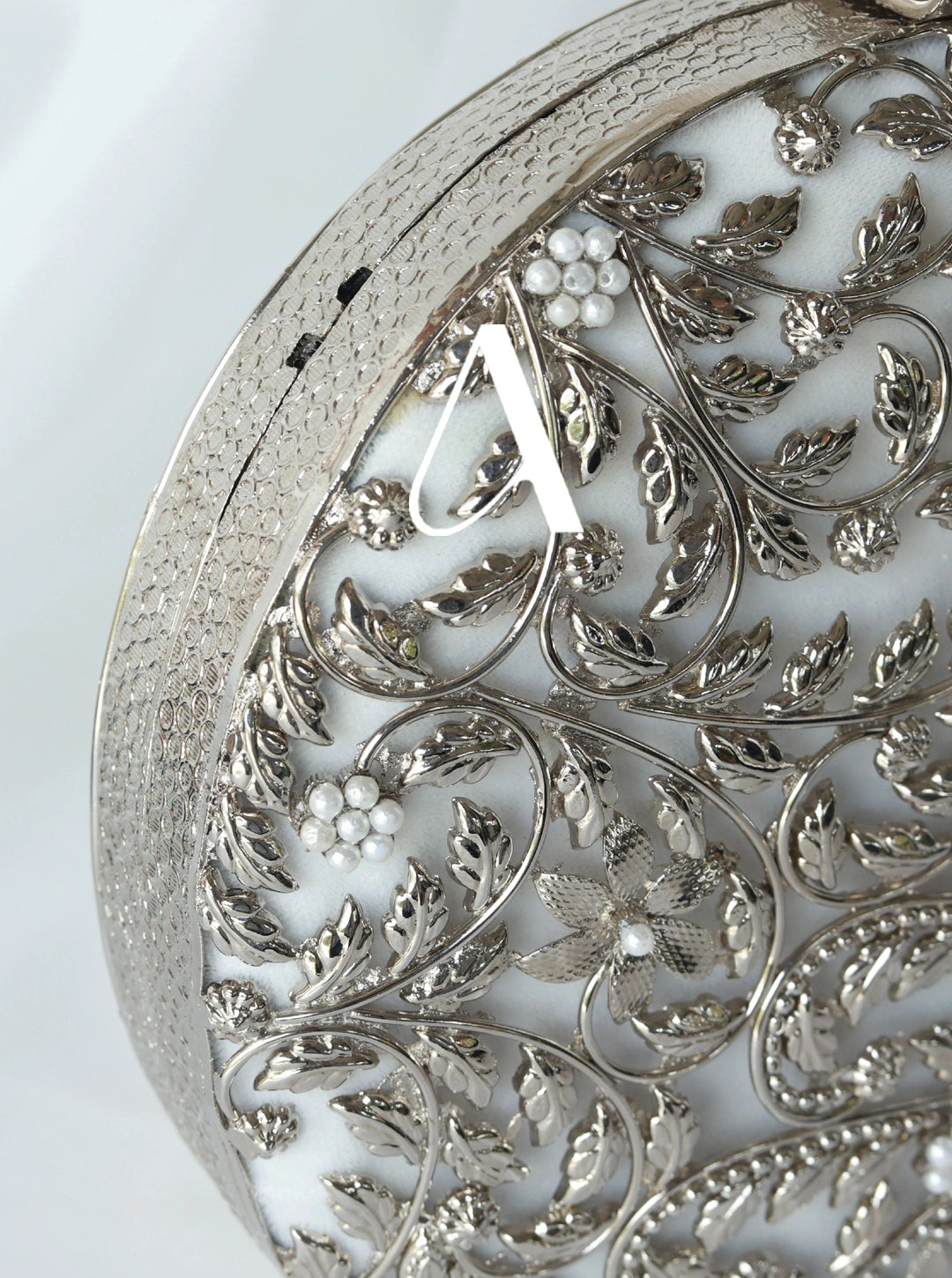 Decorative silver object with intricate leaf patterns and a central letter 'A'.