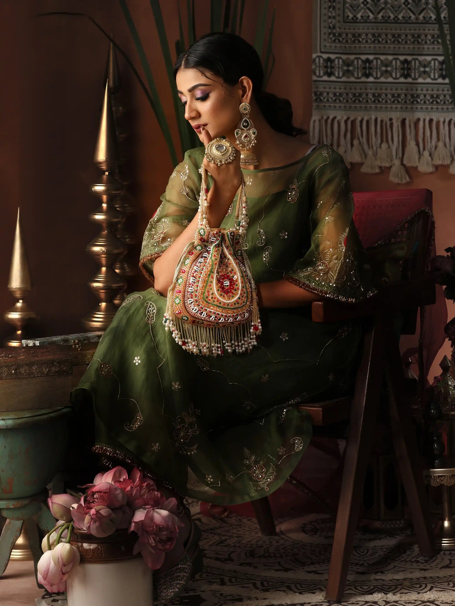 Woman in traditional green outfit with jewelry holding a decorative bag in an ornate setting.