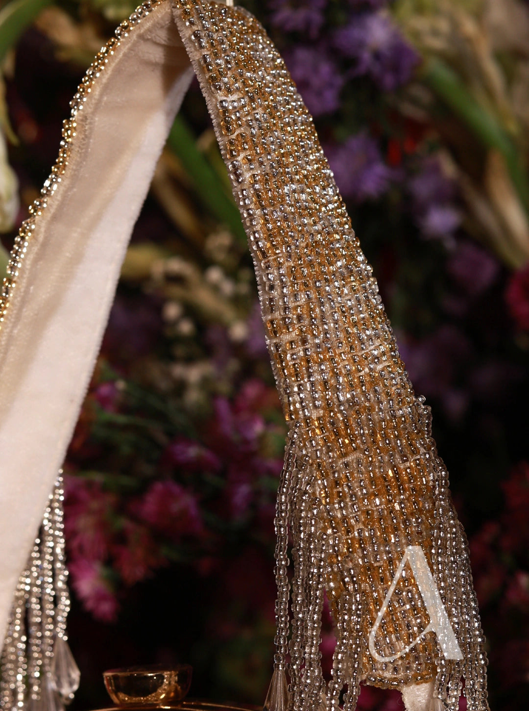 Gold beaded shoe with decorative strap featuring a letter 'A' against a blurred floral background